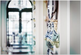 Blau-weiße Porzellanvasen mit Blumenmotiven auf vergoldeten Wandkonsolen in barockem Schlossinterieur, großes Teller-Exponat darüber, unscharfes Rundbogenfenster mit Kronleuchter im Hintergrund.