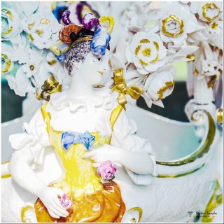 Detailaufnahme einer barocken Meissener Porzellanfigur: Dame im gelb-weißen Rüschenkleid mit blauer Schleife, buntem Kopfschmuck, umgeben von weißen und goldenen Porzellanrosen.