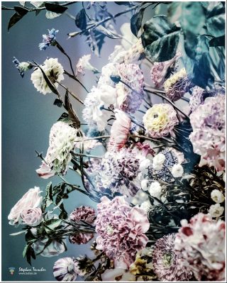 Üppiges Porzellan-Blumenarrangement mit rosa, weißen und lila Nelken, Rosen und Blättern aus bemaltem Porzellan, Spiegelung auf glänzender Fläche, blaugrauer Hintergrund.
