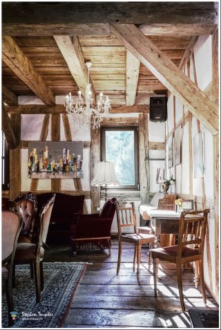 Rustikales Fachwerkinterieur mit freiliegenden Holzbalken, Kristallleuchter, antiken Samtsesseln und Holzstühlen, modernem Stadtgemälde, Orientteppich und Dielenboden im Sonnenlicht.