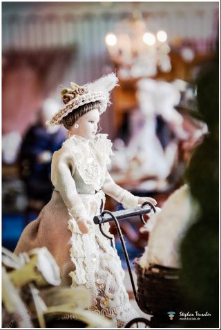 Dieses Foto ist als kostenloser Download (ohne Rand und Signatur) im Originalformat oder als Print zum Kauf/Mieten in meinem Shop auf <a href="https://butlaix.de/produkt/fotografie-puppenhaus-no-3/" target="_blank" rel="noreferrer noopener" >BUTLAIX_media</a> verfügbar. Viktorianische Porzellanpuppe in cremefarbenem Spitzenkleid und Hutschmuck an einem Miniatur-Kinderwagen, weitere unscharfe Puppen und Kronleuchter im Hintergrund.