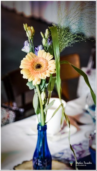 Blaue Glasvase mit Blumenstrauß: pfirsichfarbene Gerbera, lila Lisianthus-Knospen und Ziergräser, auf gedecktem Tisch mit weißer Tischdecke, unscharfer Innenraum im Hintergrund.