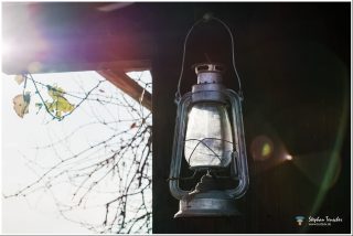 Dieses Foto ist als kostenloser Download (ohne Rand und Signatur) im Originalformat oder als Print zum Kauf/Mieten in meinem Shop auf <a href="https://butlaix.de/produkt/foto-laterne-im-sonnenlicht-als-poster-leinwand-bild-im-rahmen/" target="_blank" rel="noreferrer noopener" >BUTLAIX_media</a> verfügbar. Alte Petroleumlampe aus Metall hängt an einem Haken vor dunklem Hintergrund, mit Gegenlicht und Lens Flare, im Hintergrund kahle Äste und herbstliche Blätter.