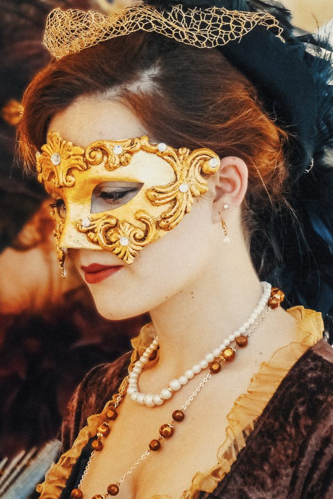 Frau mit goldener venezianischer Barock-Augenmaske, goldener Netzhaube, Perlen- und Bernsteinketten, braunem Samtkleid mit Goldbesatz. Seitenprofil, Blick nach unten.