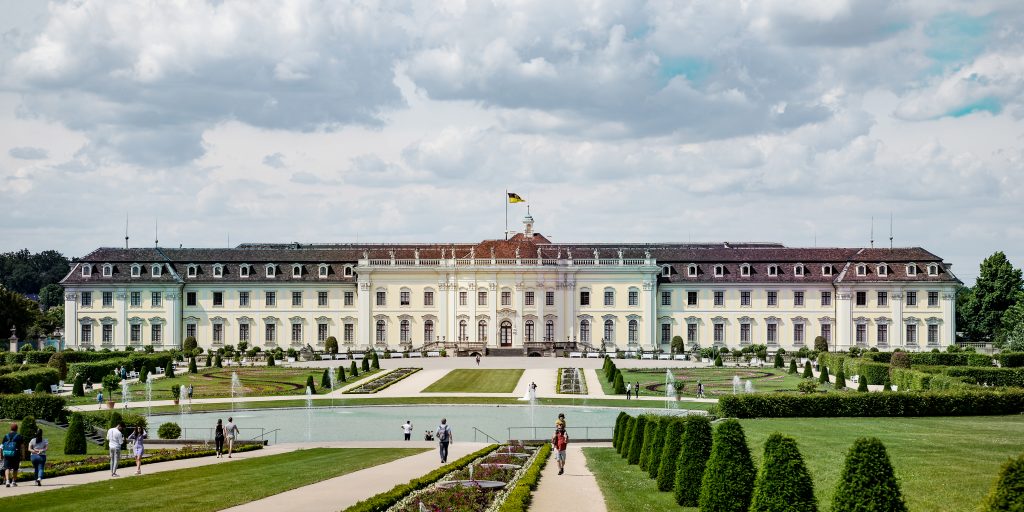 Barockschloss Ludwigsburg mit symmetrischer Fassade, davor formaler Schlossgarten mit geometrischen Rasenflächen, Wasserbecken, Springbrunnen und Spazierwegen. Besucher flanieren durch die gepflegte Gartenanlage unter bewölktem Himmel.