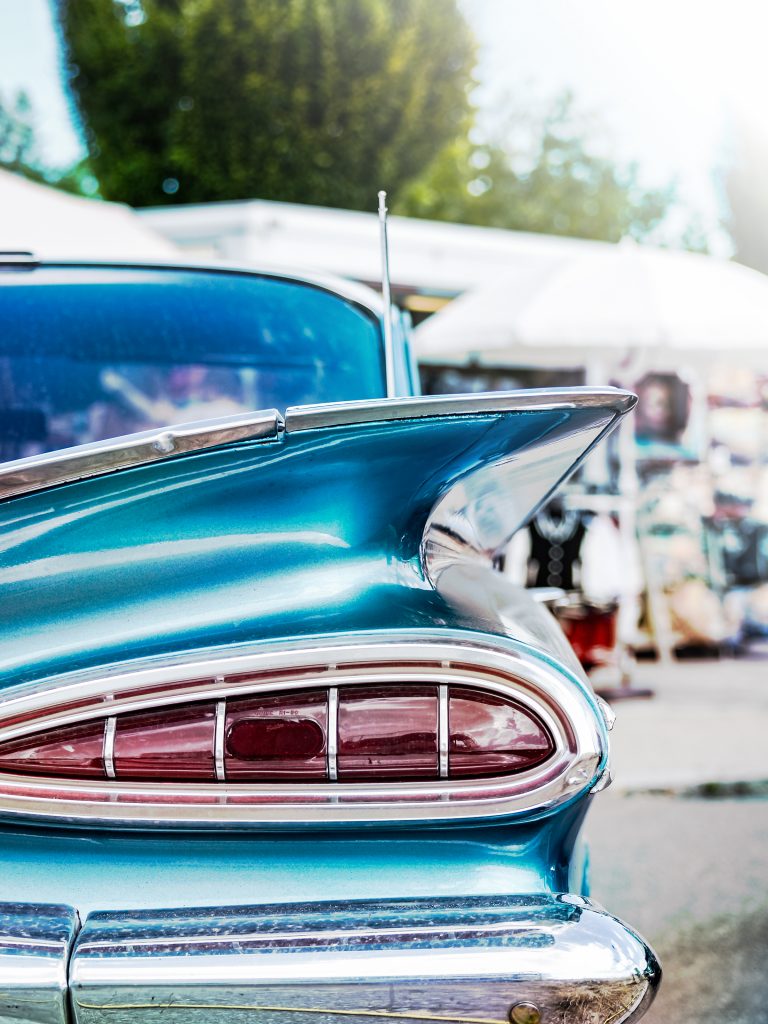 Detailaufnahme der Heckflosse eines türkisfarbenen amerikanischen Oldtimers der späten 1950er Jahre mit charakteristischem Chrom-Rücklicht, im unscharfen Hintergrund ein Veranstaltungsgelände.