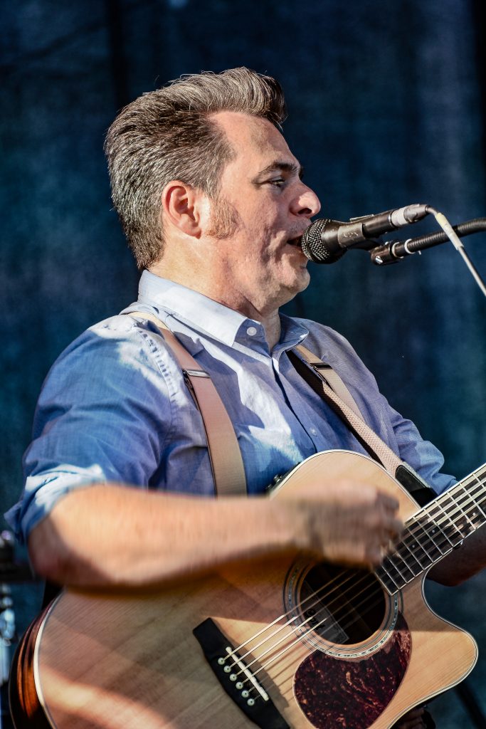 Männlicher Musiker mit kurzen braunen Haaren singt in ein Mikrofon und spielt Akustikgitarre. Er trägt ein hellblaues Hemd und braune Hosenträger. Dunkler Bühnenhintergrund.