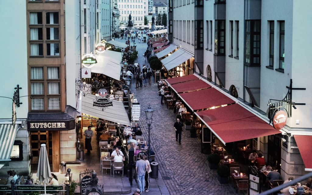 Abendliche Fußgängerzone in deutscher Altstadt mit Kopfsteinpflaster. Restaurants mit Außenterrassen unter roten und weißen Markisen säumen beide Seiten. Gäste sitzen an beleuchteten Tischen, Passanten flanieren. Links das Restaurant "Kutscher", im Hintergrund ein Platz mit Denkmal.