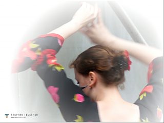 Tanzende Frau von hinten, hochgestecktes Haar mit roter Blüte, schwarzes Blumenkleid mit roten Rosen, Arme ausgestreckt, Bewegungsunschärfe, weich ausgeblendete Bildränder.