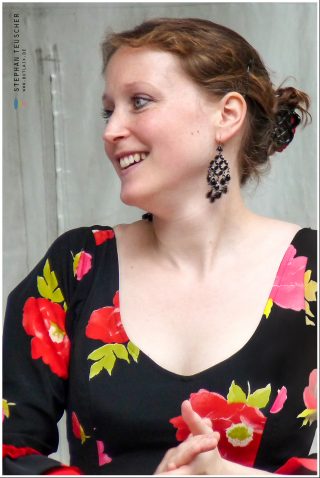 Lächelnde Frau im Halbprofil, hochgestecktes Haar, auffällige dunkle Hängeohrringe, schwarzes Flamenco-Kleid mit roten und rosafarbenen Blüten, vor grauem Hintergrund.
