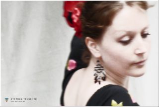 Frau im Seitenprofil, Blick nach unten, hochgestecktes Haar mit roter Blüte, dunkle Hängeohrringe, schwarzes Blumenkleid, weiche Unschärfe, heller grauer Hintergrund.
