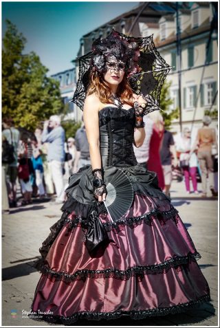 Frau in Gothic-Viktorianischem Kostüm: schwarz-bordeauxfarbenes Ballkleid mit Korsett, schwarzer Spitzenschirm, filigrane Augenmaske und Fächer. Outdoor-Auftritt auf belebtem Stadtplatz.
