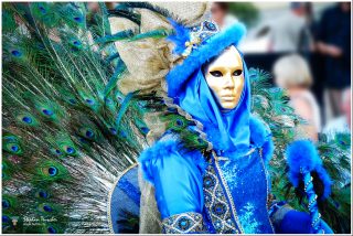 Venezianische Kostümfigur in leuchtendem Blau mit goldener Vollmaske, Pfauenfedern-Rückenschmuck, Pailletten-Gewand und Pelzbesatz. Eindrucksvolles Pfauenmotiv-Kostüm, Bokeh-Hintergrund.