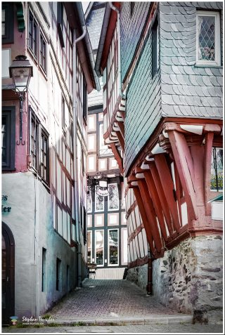Dieses Foto ist als kostenloser Download (ohne Rand und Signatur) im Originalformat oder als Print zum Kauf/Mieten in meinem Shop auf <a href="https://butlaix.de/produkt/fotografie-altstadt-von-limburg-an-der-lahn-no-1/" target="_blank" rel="noreferrer noopener" >BUTLAIX_media</a> verfügbar. Enge Gasse mit historischen Fachwerkhäusern in einer deutschen Altstadt. Bunte Gebäude mit weißem Putz, roten und grünen Holzbalken, Schieferdächern und Kopfsteinpflaster. Alte Laternen und Ladenschilder säumen den Weg.