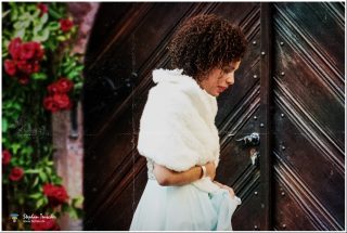 Frau mit lockigem braunem Haar im Profil vor einer dunklen Holztür, weißer Pelzstola über hellblauem Kleid, links rote Rosen an einer Mauer.