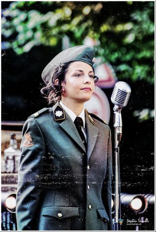 Dieses Foto ist als kostenloser Download (ohne Rand und Signatur) im Originalformat oder als Print zum Kauf/Mieten in meinem Shop auf <a href="https://butlaix.de/produkt/fotografie-vintage-swing-performance-no-1/" target="_blank" rel="noreferrer noopener" >BUTLAIX_media</a> verfügbar. Junge Frau in grauer Militäruniform mit Schirmmütze steht vor einem Vintage-Mikrofon im Freien. Das Foto hat einen körnigen, nostalgischen Filter im Stil der 1940er Jahre.