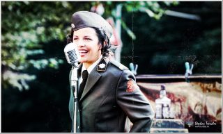 Dieses Foto ist als kostenloser Download (ohne Rand und Signatur) im Originalformat oder als Print zum Kauf/Mieten in meinem Shop auf <a href="https://butlaix.de/produkt/fotografie-vintage-swing-performance-no-6/" target="_blank" rel="noreferrer noopener" >BUTLAIX_media</a> verfügbar. Lächelnde Sängerin in niederländischer Retro-Militäruniform mit Barettmütze seitlich am Vintage-Chromständermikrofon, Bäume und Requisiten unscharf im Hintergrund, Kratzer-Effekt.
