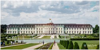 Barockschloss Ludwigsburg mit symmetrischer Fassade, davor formaler Schlossgarten mit geometrischen Rasenflächen, Wasserbecken, Springbrunnen und Spazierwegen. Besucher flanieren durch die gepflegte Gartenanlage unter bewölktem Himmel.