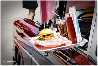 Dieses Foto ist als kostenloser Download (ohne Rand und Signatur) im Originalformat oder als Print zum Kauf/Mieten in meinem Shop auf <a href="https://butlaix.de/produkt/fotografie-american-50s-lifestyle/" target="_blank" rel="noreferrer noopener" >BUTLAIX_media</a> verfügbar. Tablett mit Burger, Pommes und Cola an der Autotür eines roten Oldtimers befestigt, im Stil eines amerikanischen 1950er-Jahre Drive-in-Restaurants, kleine US-Flagge im Burger.