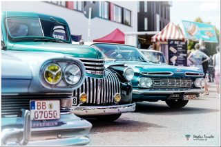Dieses Foto ist als kostenloser Download (ohne Rand und Signatur) im Originalformat oder als Print zum Kauf/Mieten in meinem Shop auf <a href="https://butlaix.de/produkt/fotografie-oldtimer-no-8/" target="_blank" rel="noreferrer noopener" >BUTLAIX_media</a> verfügbar. Drei klassische amerikanische Oldtimer in Türkis- und Grüntönen auf einem Autotreffen. Im Vordergrund ein Chevrolet mit markanter Chromfront und gelben Scheinwerfern. Im Hintergrund Imbissstände und Besucher.