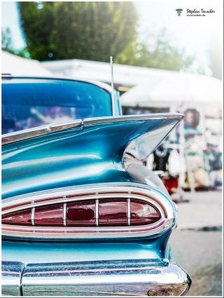 Dieses Foto ist als kostenloser Download (ohne Rand und Signatur) im Originalformat oder als Print zum Kauf/Mieten in meinem Shop auf <a href="https://butlaix.de/produkt/fotografie-oldtimer-no-10/" target="_blank" rel="noreferrer noopener" >BUTLAIX_media</a> verfügbar. Detailaufnahme der Heckflosse eines türkisfarbenen amerikanischen Oldtimers der späten 1950er Jahre mit charakteristischem Chrom-Rücklicht, im unscharfen Hintergrund ein Veranstaltungsgelände.