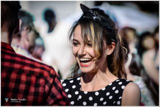 Lachende junge Frau im schwarz-weißen Polka-Dot-Kleid mit schwarzem Bandana-Haarband, umgeben von Menschen bei einem Outdoor-Festival. Retro-Stil, Bokeh-Hintergrund.