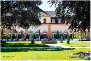 Dieses Foto ist als kostenloser Download (ohne Rand und Signatur) im Originalformat oder als Print zum Kauf/Mieten in meinem Shop auf <a href="https://butlaix.de/produkt/fotografie-schloss-weilburg-no-11/" target="_blank" rel="noreferrer noopener" >BUTLAIX_media</a> verfügbar. Das Foto zeigt eine elegante Barockarchitektur mit symmetrischen Fenstern und einem gepflegten Garten. Statuen, ein Brunnen und Palmen zieren den Hof, während Schatten großer Bäume eine malerische Atmosphäre schaffen. Stilvolle Sonnenschirme ergänzen das Ambiente.