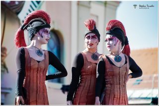 Drei Performerinnen in identischen Kostümen: rostfarbene Kleider, schwarze Helme mit roten Federbüschen, dramatisches Bühnen-Make-up in Rot-Schwarz und runde Anhänger. Outdoor-Auftritt bei Tageslicht.