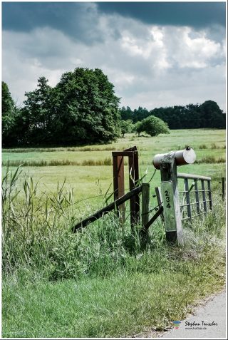 Dieses Foto ist als kostenloser Download (ohne Rand und Signatur) im Originalformat oder als Print zum Kauf/Mieten in meinem Shop auf <a href="https://butlaix.de/produkt/fotografie-post-fuer-234/" target="_blank" rel="noreferrer noopener" >BUTLAIX_media</a> verfügbar. Ländliche Szene mit verwittertem Holzzauntor und Briefkasten mit Nummer 234 am Straßenrand. Dahinter grüne Wiese, Laubbäume und bewölkter Himmel mit dramatischen Wolken.