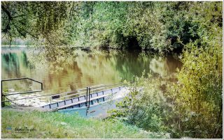 Dieses Foto ist als kostenloser Download (ohne Rand und Signatur) im Originalformat oder als Print zum Kauf/Mieten in meinem Shop auf <a href="https://butlaix.de/produkt/fotografie-an-der-lahn-no-4/" target="_blank" rel="noreferrer noopener" >BUTLAIX_media</a> verfügbar. Ein idyllischer Flussabschnitt, umgeben von dichtem, grünem Laub. Im Vordergrund liegt ein kleines, metallisches Boot am Ufer, teils verdeckt von Pflanzen. Die ruhige Wasseroberfläche reflektiert die Bäume.