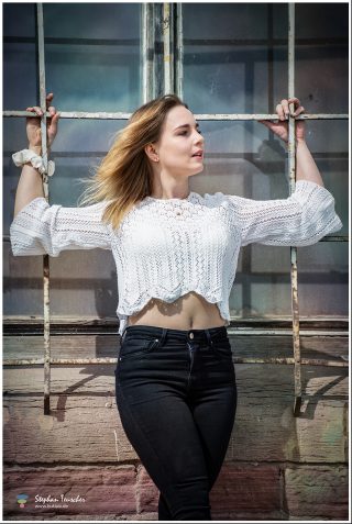 Junge Frau in weißem Häkelpullover und schwarzer Jeans hält sich an einem verwitterten Industriefenster fest und blickt seitlich. Wehende Haare, freigelegter Bauch, Backsteinwand im Hintergrund.