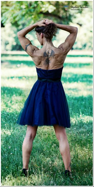 Rückansicht einer muskulösen Frau in dunkelblauem, rückenfreiem Tüllkleid auf einer Wiese. Sie hält ihr hochgestecktes Haar mit beiden Händen. Auf ihrem Rücken ist ein großes Tattoo mit Drachenflügeln sichtbar. Im Hintergrund unscharfe Bäume und Gras.