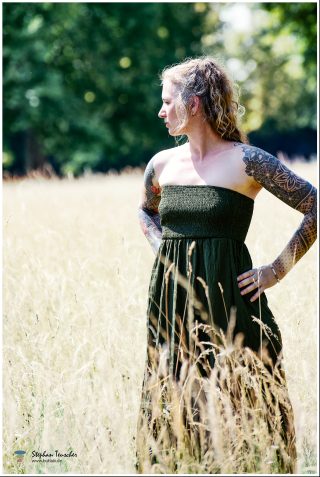 Frau mit lockigem blondem Haar im Profil, trägt ein schulterfreies dunkelgrünes Kleid. Beide Arme sind mit detaillierten geometrischen und floralen Tattoos bedeckt. Sie steht in einem sonnigen Feld mit hohem, trockenem Gras, im Hintergrund grüne Bäume.