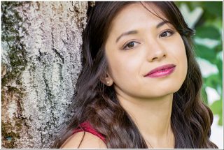Porträt einer jungen Frau mit langen braunen Haaren und braunen Augen, die an einem Baumstamm lehnt. Sie trägt pinken Lippenstift, kleine Ohrstecker und ein rotes Top. Der Hintergrund zeigt unscharfes grünes Laub.