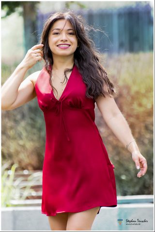 Junge Frau mit langem, welligem braunem Haar in einem roten Satin-Minikleid mit V-Ausschnitt und Schleife. Sie lächelt, trägt pink Lippenstift, eine Halskette und hat ein kleines Tattoo am Handgelenk. Unscharfer Garten-Hintergrund bei Tageslicht.