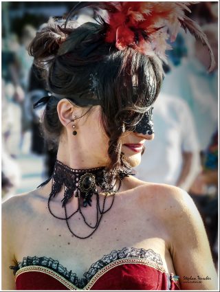 Frau im Vintage-Stil mit hochgestecktem dunklem Haar, geschmückt mit roten und schwarzen Federn. Sie trägt eine schwarze Spitzenmaske, ein ornamentales schwarzes Collier mit Ketten und ein rotes Korsett mit Spitzenbesatz. Seitliches Profil, lächelnd.