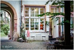 Historischer Innenhof mit Kopfsteinpflaster, großem Rundbogen-Durchgang und weißer Hausfassade. Vor einem Schaufenster stehen weiße Metallstühle und ein Tisch. Grüne Pflanzen und Efeu ranken an den altrosa gestrichenen Wänden. Gemütliche, verwunschene Atmosphäre.