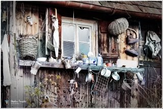 Verwitterte Holzhütte mit Fenster, davor ein Regal voller Gegenstände: Emailletassen, blau-weiße Keramikkrüge, Moka-Kanne, Weidenkörbe, Regenschirm, Wanderschuhe, Rucksack und diverses Küchengeschirr. Kupferne Dachrinne, rankende Pflanzen.