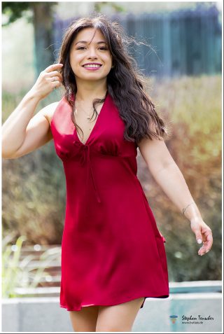 Junge Frau mit langem, welligem braunem Haar in einem roten Satin-Minikleid mit V-Ausschnitt und Schleife. Sie lächelt, trägt pink Lippenstift, eine Halskette und hat ein kleines Tattoo am Handgelenk. Unscharfer Garten-Hintergrund bei Tageslicht.